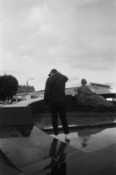 A person stands near a statue in a reflective urban park, captured in black and white.