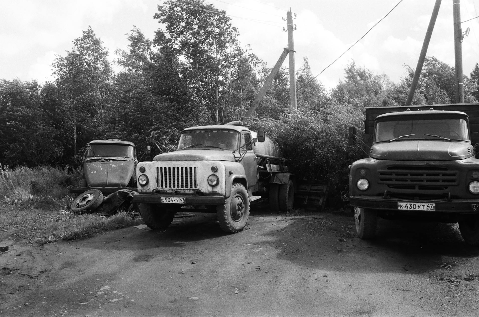 A black and white photo of abandoned vintage trucks parked outdoors, surrounded by trees.