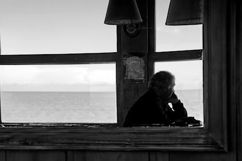 Grayscale Photo of Man Sitting by the Window Looking at the Sea