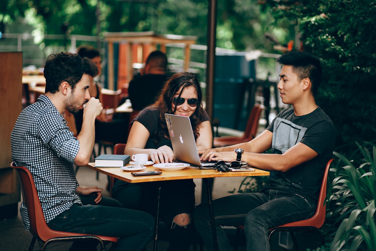 Three young professionals chatting and using a laptop at an outdoor table, highlighting casual networking.