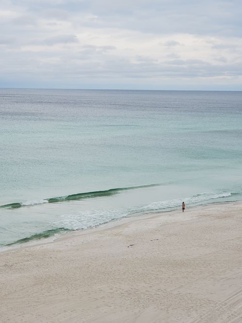 Free A woman walks alone on a tranquil Panama City Beach shoreline under a cloudy sky. Stock Photo