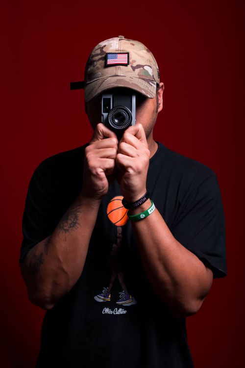 Free Man wearing a cap holds a vintage camera against a red background in a studio setting. Stock Photo