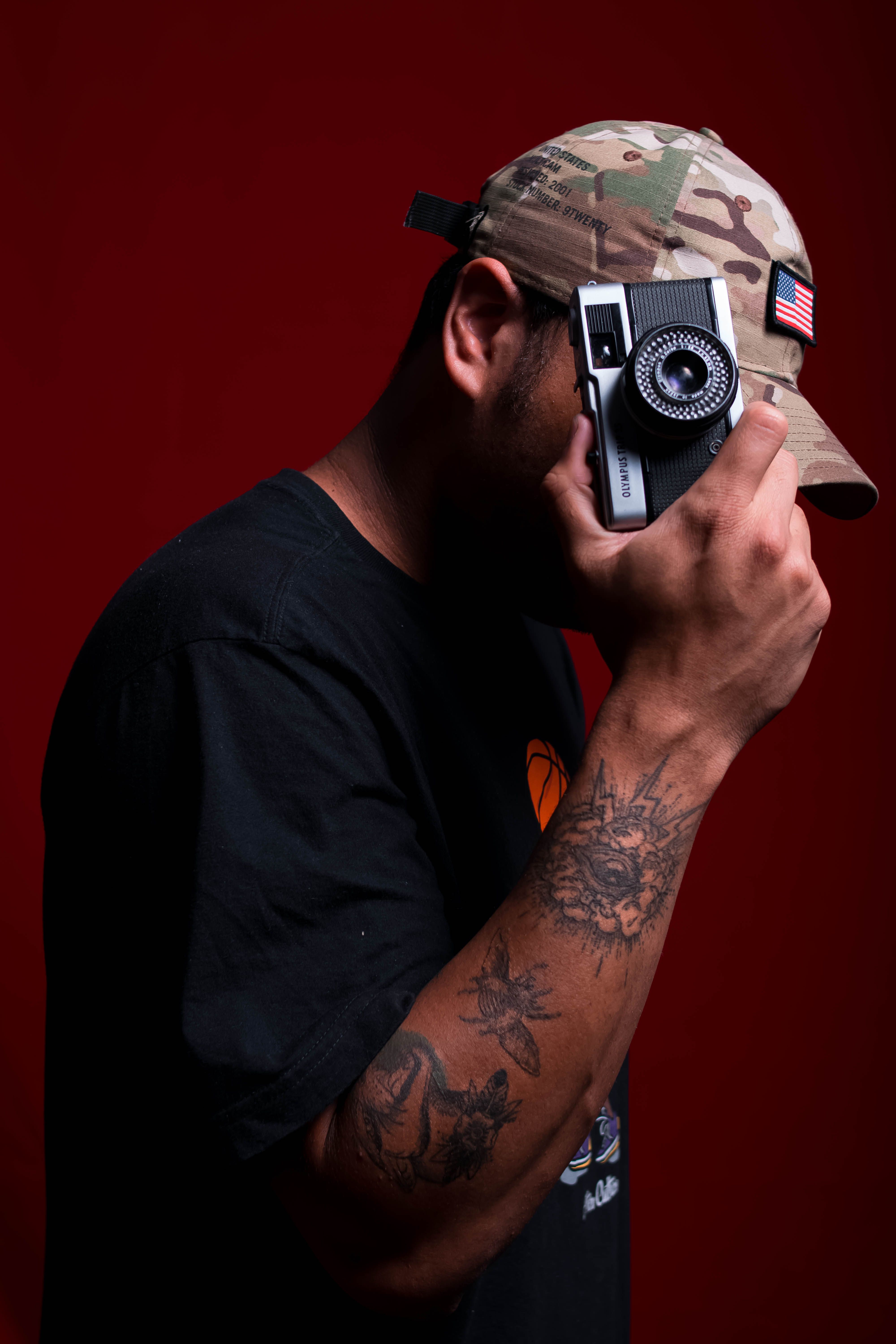 Man with arm tattoos and camouflage cap holding a vintage camera against a red backdrop.