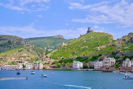 Picturesque coastal town with boats, lush hills, and historical buildings under a clear blue sky.
