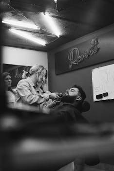 Black and white photo of a barber shaving a customer's beard, capturing salon authenticity.