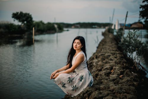 Free Woman in White and Blue Floral Dress Sitting on Rock Near Body of Water Stock Photo