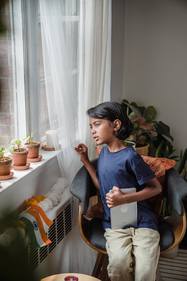 A Boy Looking Out A Window While Holding A Tablet