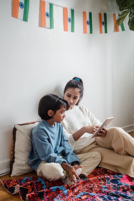 A mother and son sit cross-legged on a colorful carpet using a digital tablet indoors.