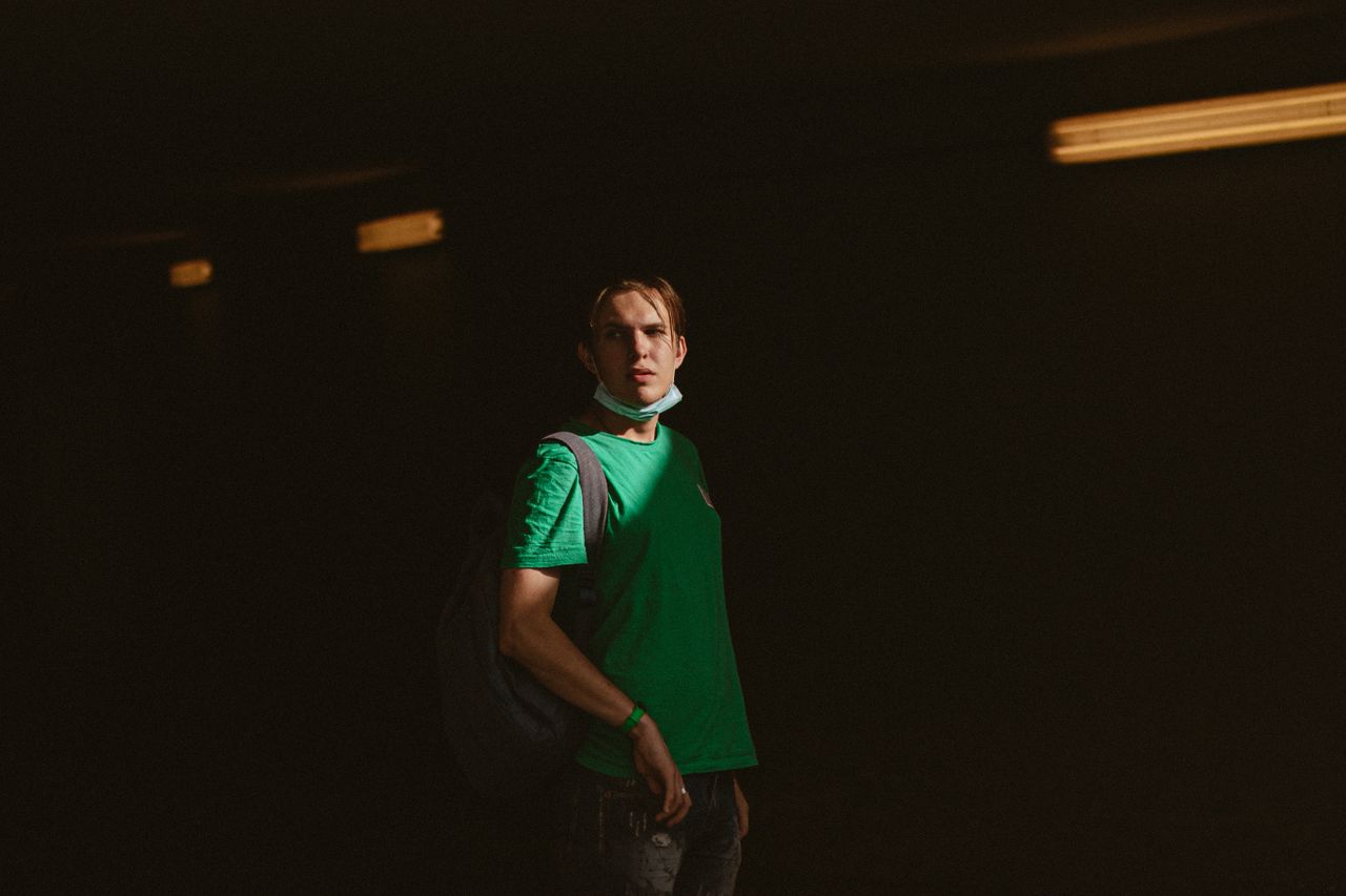 a man wearing a green shirt and backpack stands in shadow face mask lo