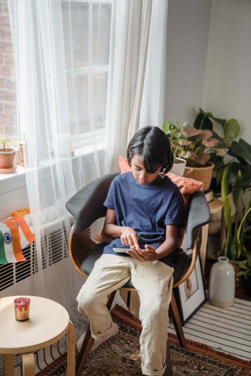 Free A young boy sits indoors using a smartphone, surrounded by plants and soft lighting. Stock Photo
