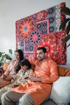 Family wearing traditional Indian clothing enjoys relaxation time on sofa with vibrant tapestry background.