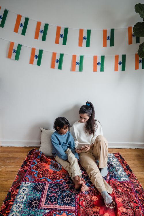 Free A mother and her son sitting on a colorful rug, using a tablet in a room with Indian flags hung as decoration. Stock Photo