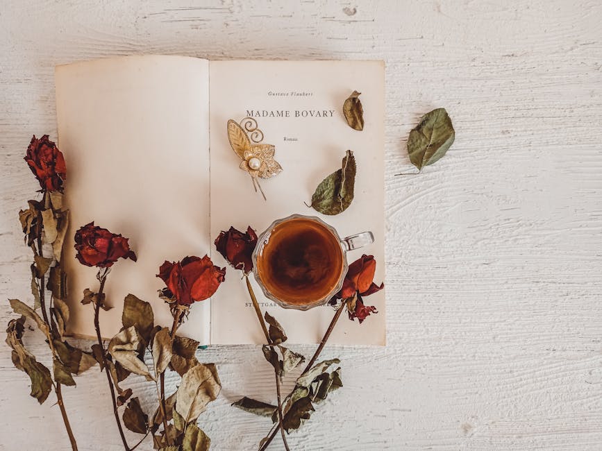 Aesthetic flat lay featuring Madame Bovary, a brooch, tea, and dried roses on a wooden surface.