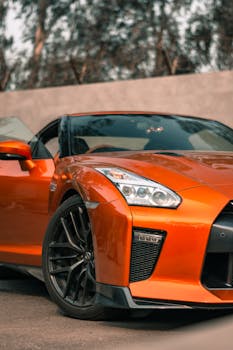 Close-up of a shiny orange sportscar, highlighting its headlight and sleek design against an outdoor backdrop.