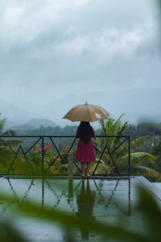 A woman stands on a terrace with an umbrella, overlooking a misty mountain view in a tropical setting.
