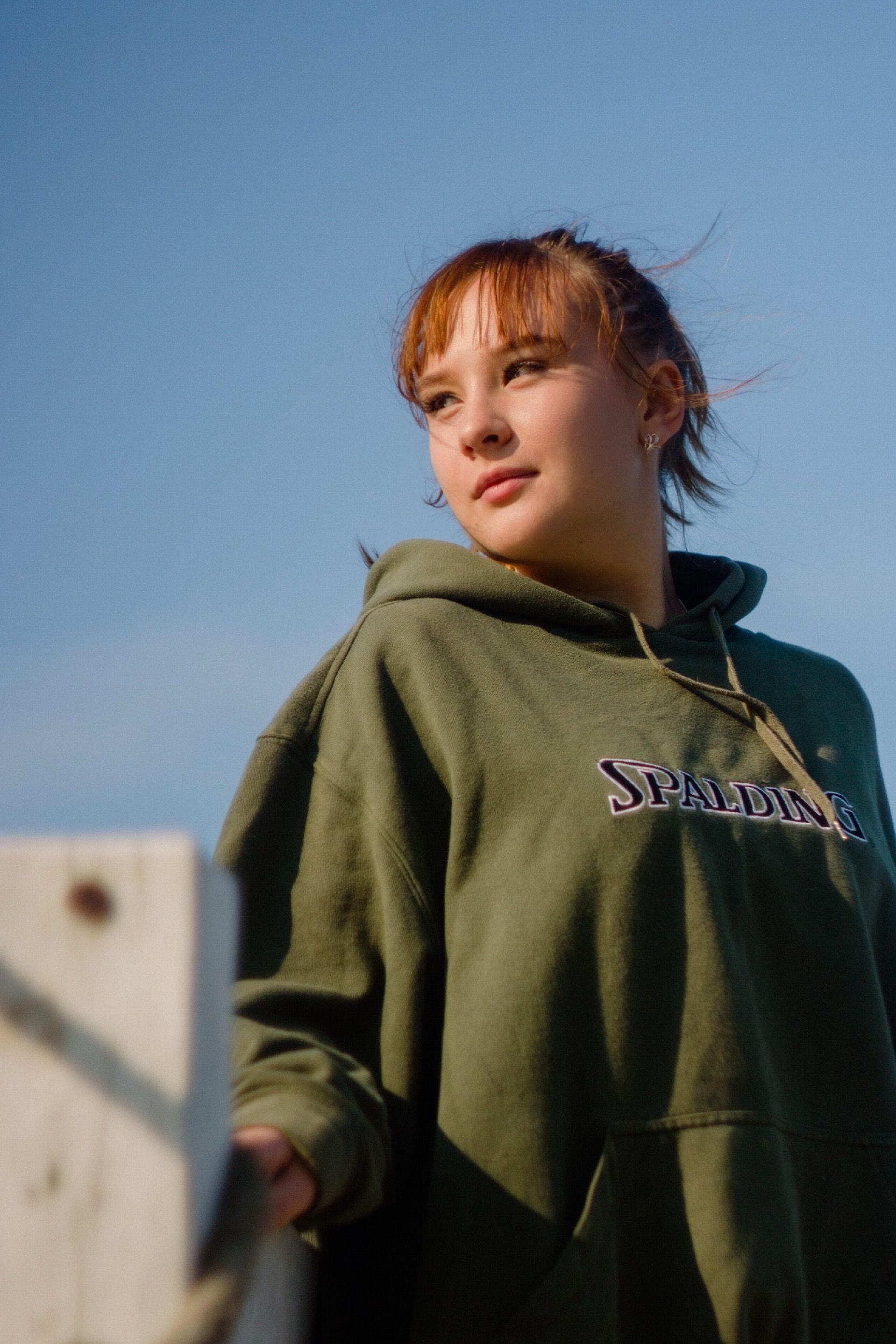 Portrait of a young woman in a green hoodie, looking away outdoors under clear sky.