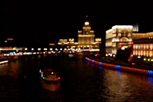 Defocused view of illuminated cityscape and river at night with boats and colorful reflections.