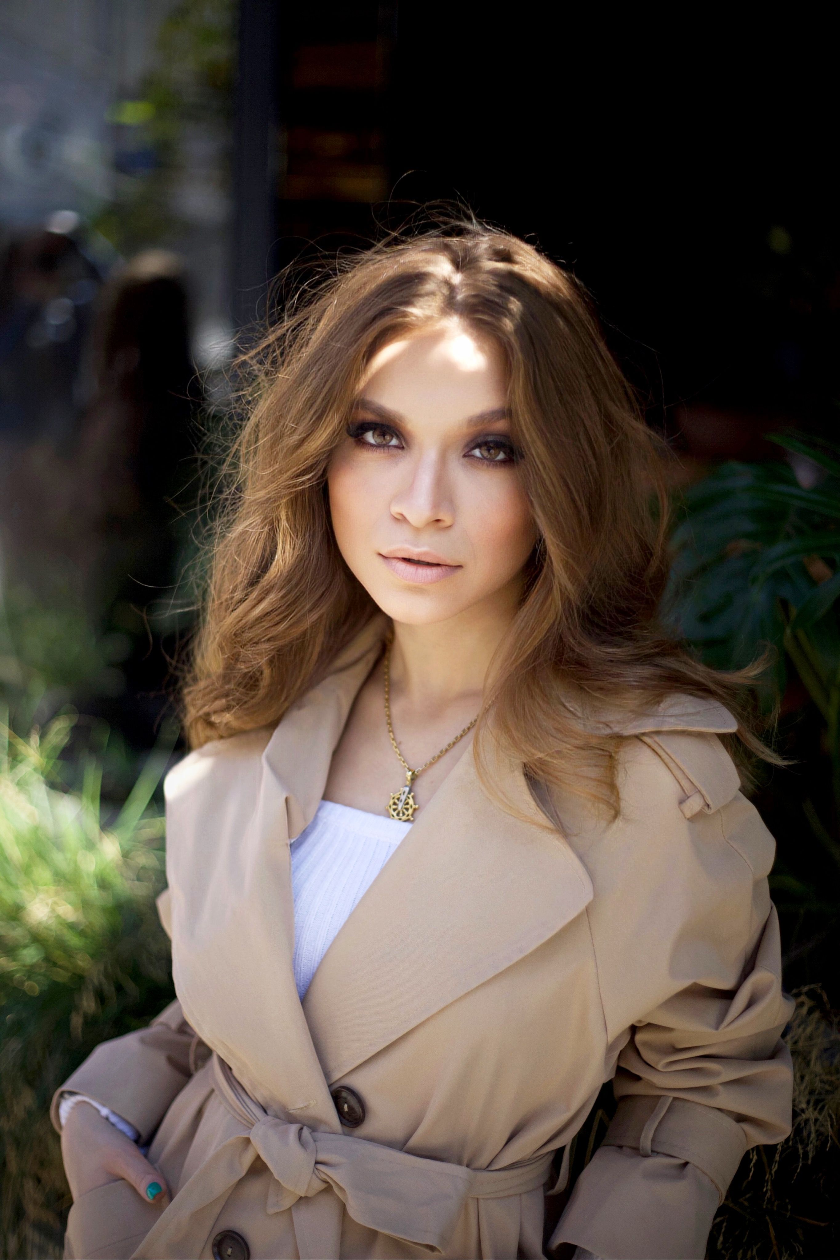 Stylish portrait of a woman in a trench coat posing elegantly outdoors in natural light.