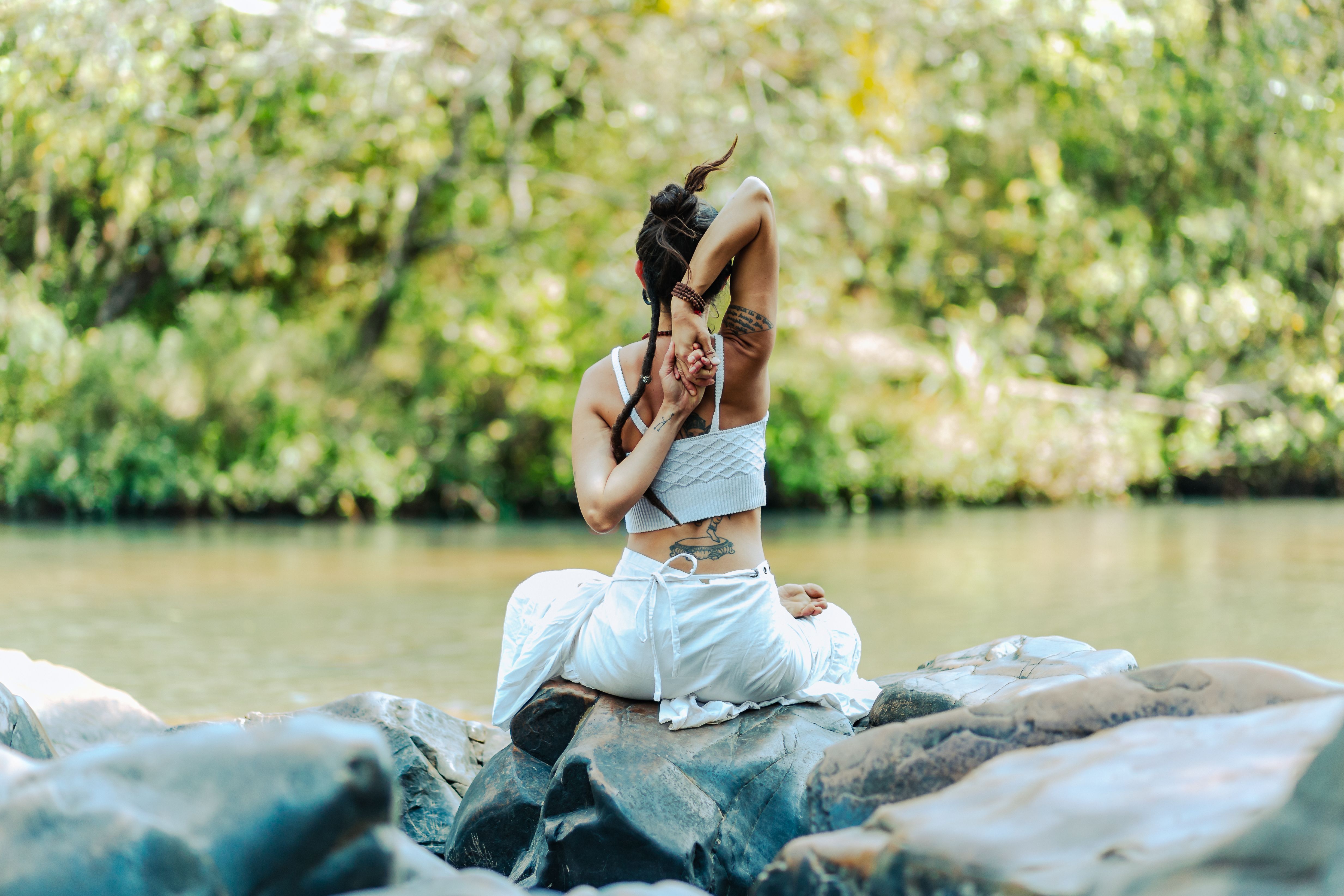 A serene scene of a woman with tattoos meditating in a yoga pose on rocks by a river.