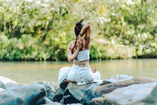 A serene scene of a woman with tattoos meditating in a yoga pose on rocks by a river.