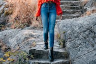 Woman in Blue Denim Jeans and Red Coat Standing on Gray Rock
