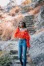 Woman in Orange Off Shoulder Shirt and Blue Denim Jeans Standing on Brown Rock