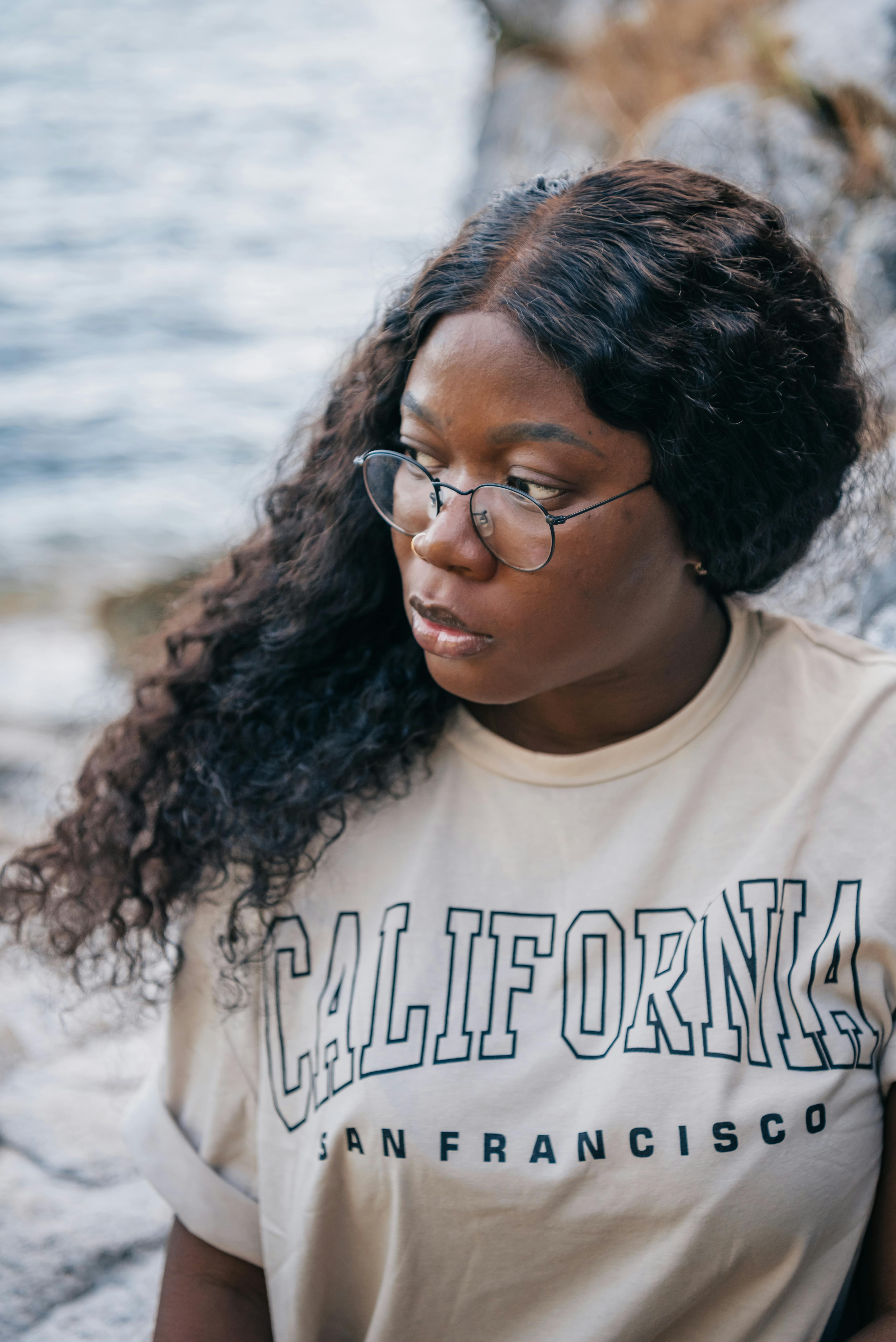 Thoughtful woman in glasses by the water, wearing a California t-shirt.