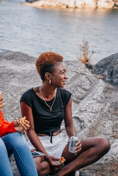 A young woman sits on a rock holding a drink by a peaceful lakeside during the day.