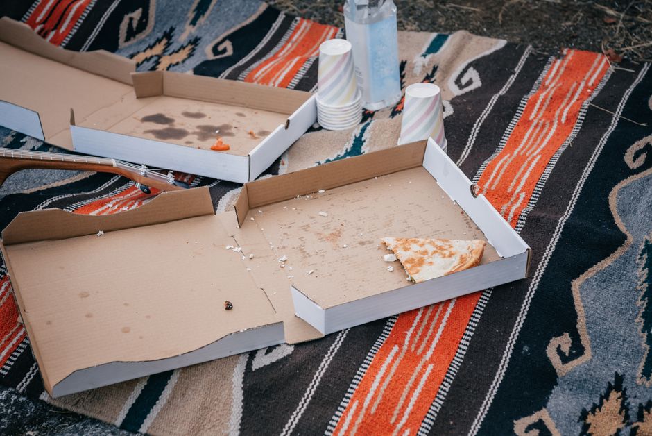 A casual picnic scene with empty pizza boxes and paper cups on a patterned blanket.