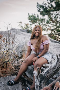 Smiling young woman in casual wear sits on a rock, surrounded by nature during a sunny day.