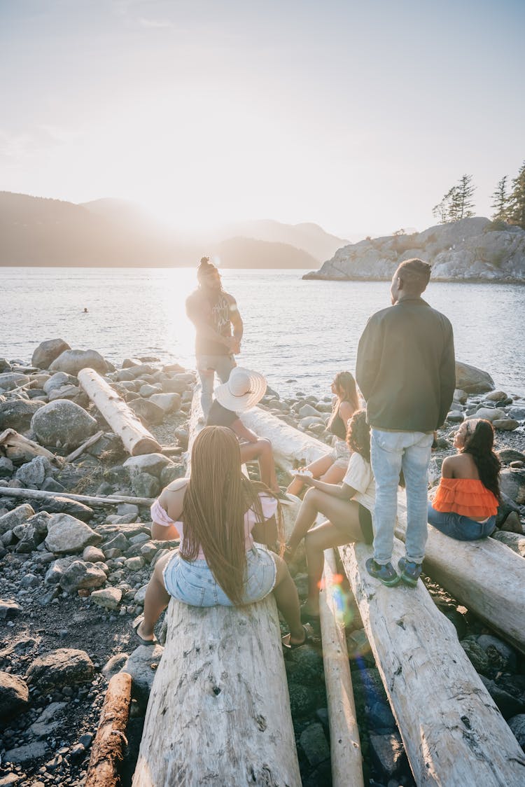 People Standing And Sitting On Wooden Logs Near A Body Of Water