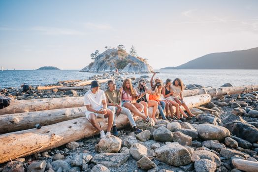 Friends relax on logs by the rocky shore, enjoying a sunny day at the beach.