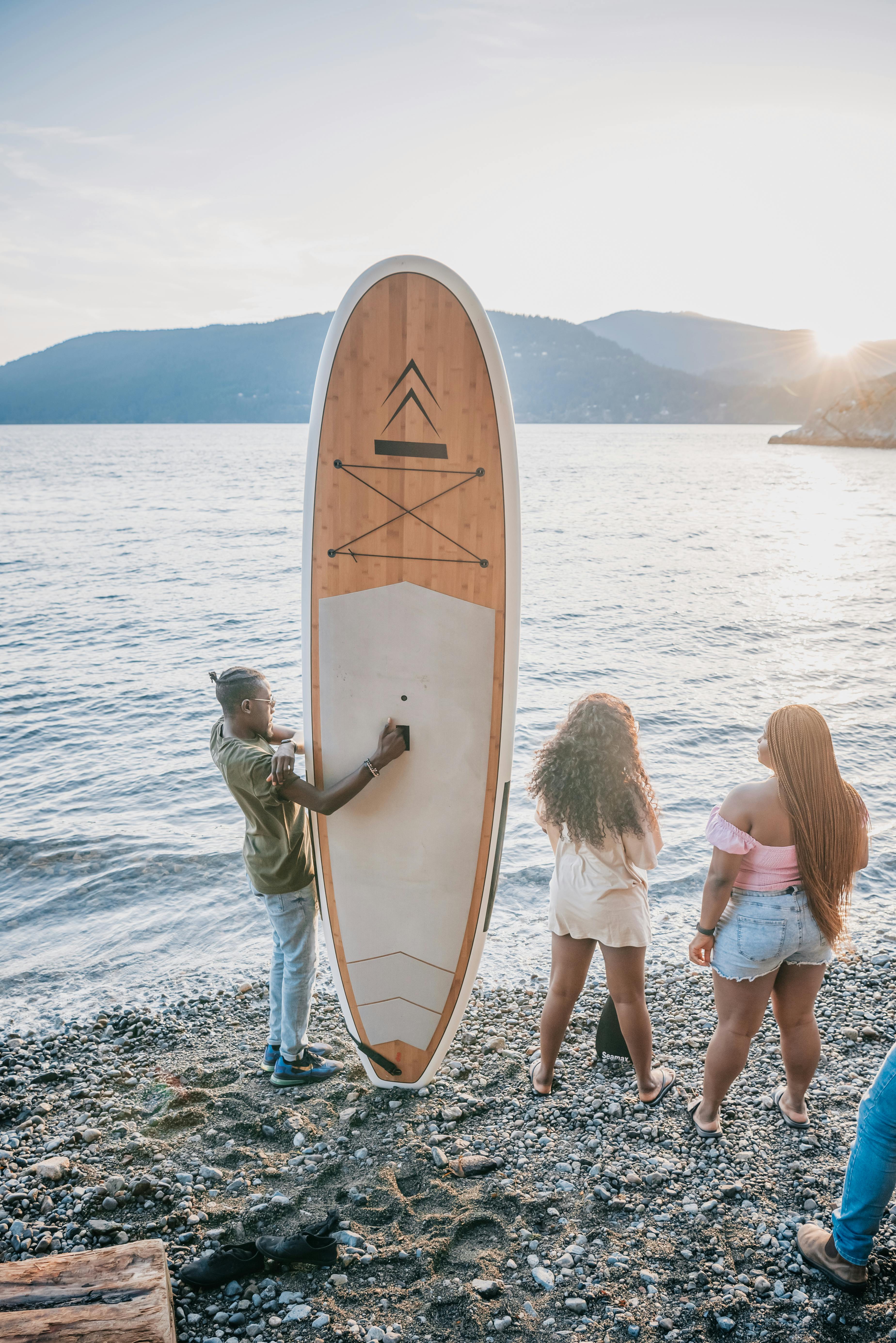 A group of adults relax by the shore with a surfboard, enjoying a warm sunset at the beach.