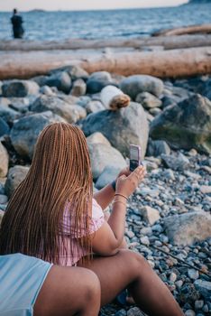 Woman with braided hair using smartphone on a rocky beach, capturing ocean scenery during summer.