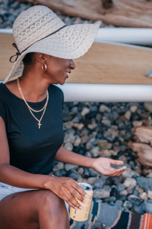 Free Black woman in a sun hat enjoys a canned beverage while sitting outdoors, exuding a relaxed vibe. Stock Photo