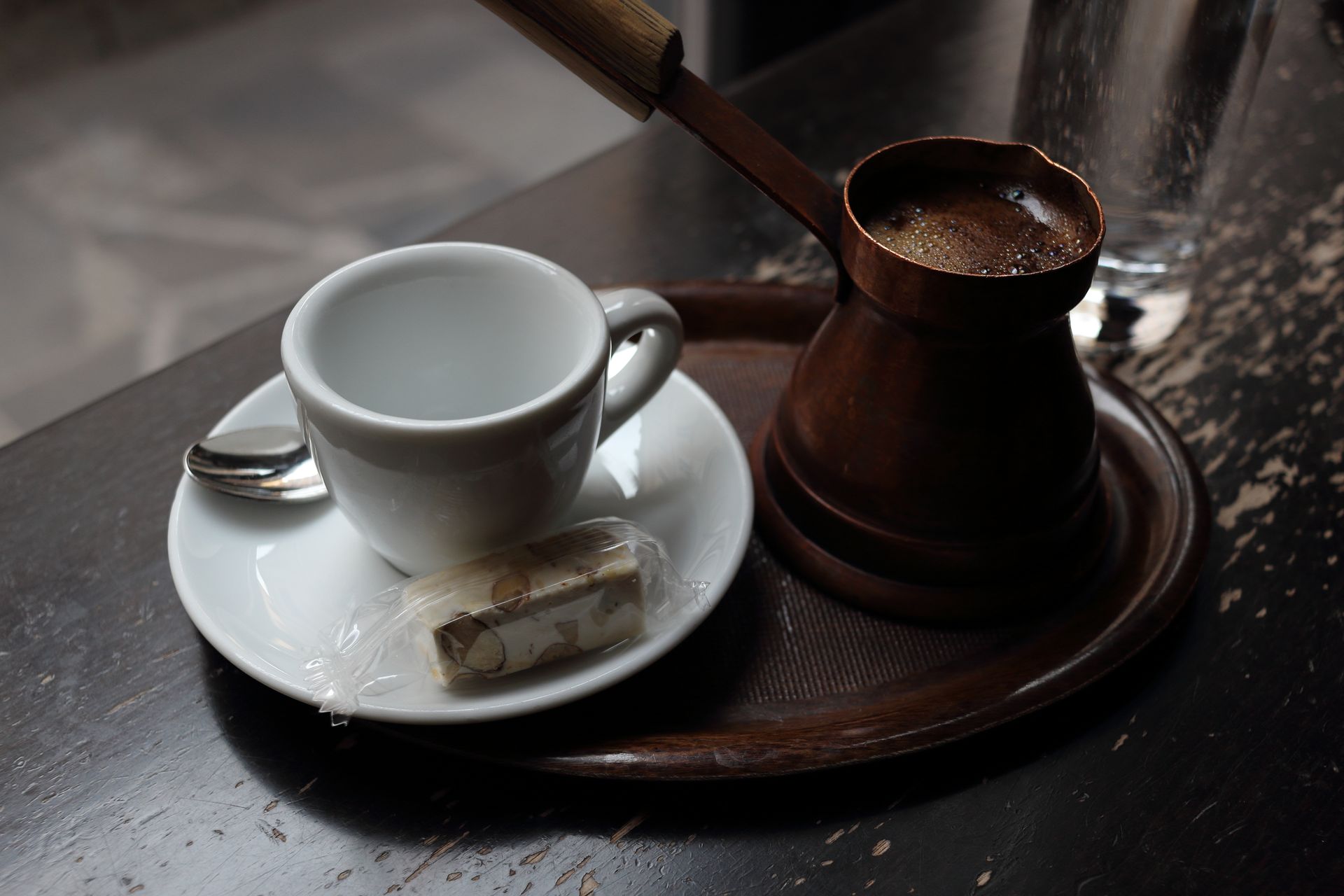 Close-up of a ceramic coffee cup, copper pot, and candy bar on a rustic table.