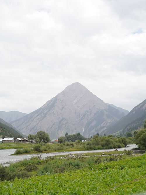 Free A towering mountain stands majestically above a lush valley with greenery and a flowing river beneath a cloudy sky. Stock Photo