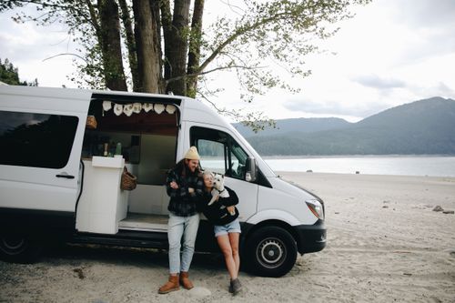 Free Couple embraces their dog by a campervan at a scenic lakeside, showcasing outdoor van life. Stock Photo