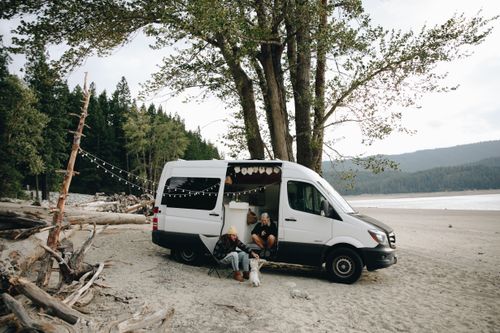Free Couple sitting by camper van on a serene beach with their dog, enjoying a peaceful outdoor escape. Stock Photo