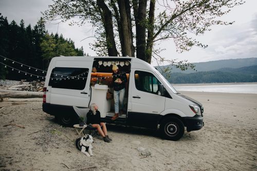Free A couple with their dog relax by a camper van on a serene beach, embracing outdoor lifestyle. Stock Photo