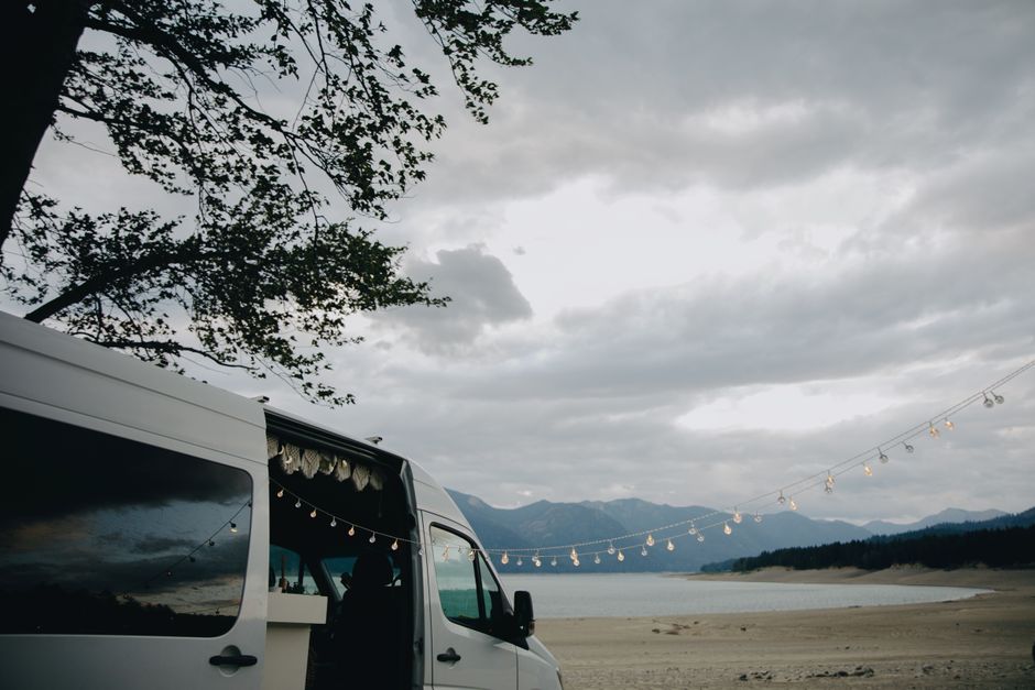 A campervan parked at a beach with string lights and a serene mountain backdrop at sunset.