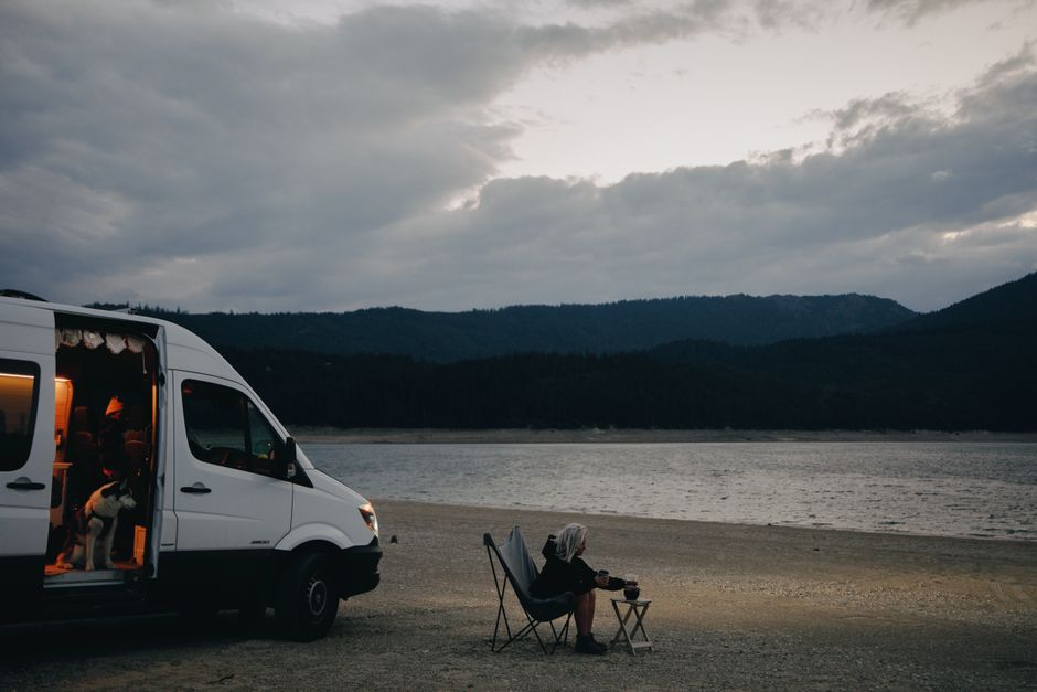 A woman relaxes by a lakeside, seated next to a campervan with a dog and folding chair under cloudy skies.