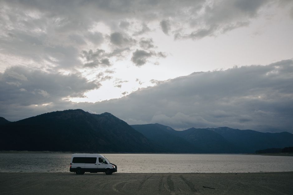 A campervan parked on a beach with mountains under a cloudy dawn sky.