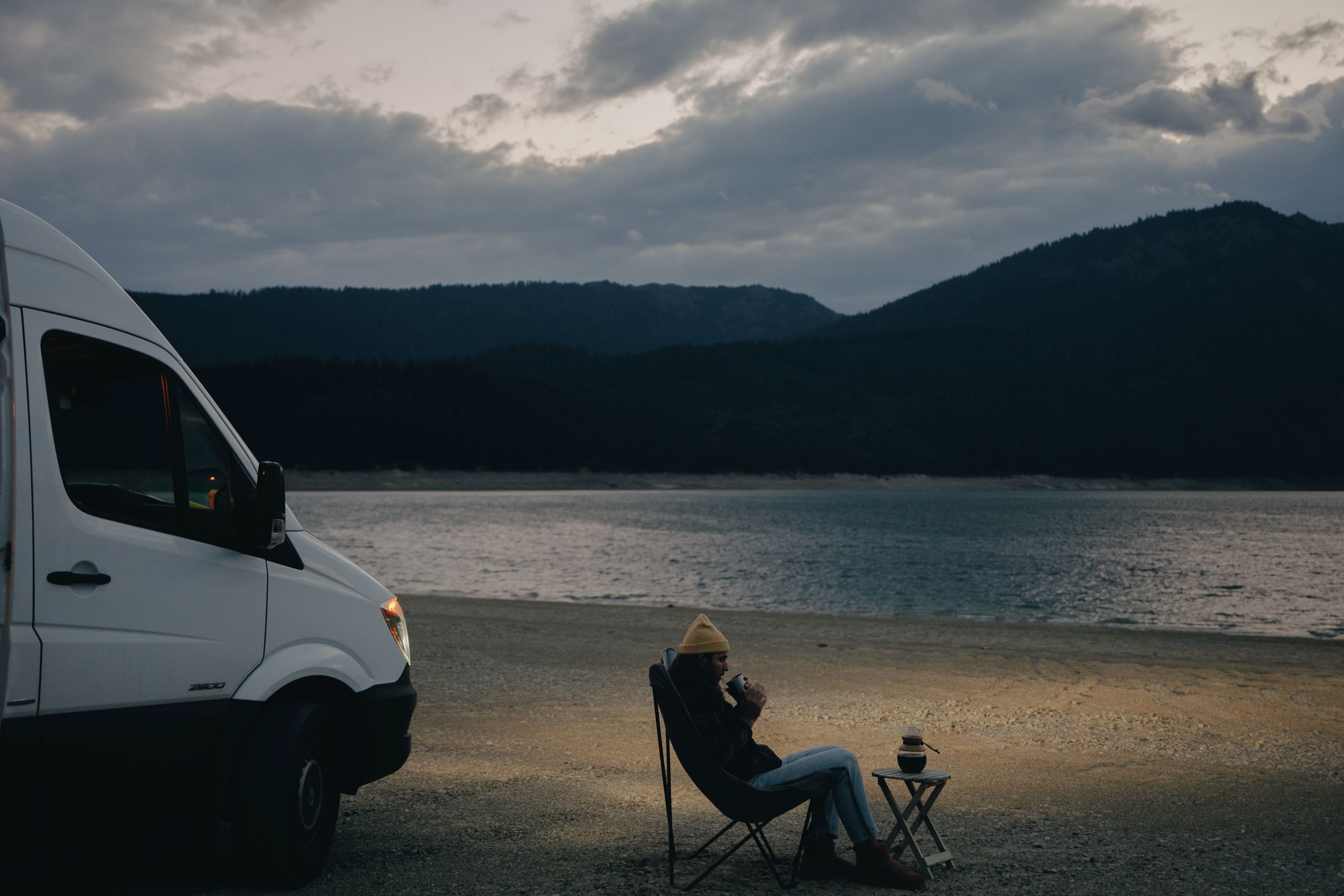 A person enjoys a peaceful evening by the lake with a camper van and mountains in the background.