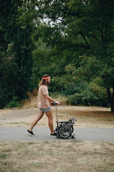 Casually dressed man pushing a folding bike on a park path surrounded by greenery.