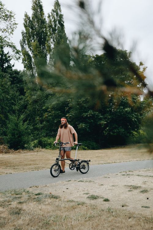 Free A man strolls in a park, leading a foldable bicycle along a path. Stock Photo
