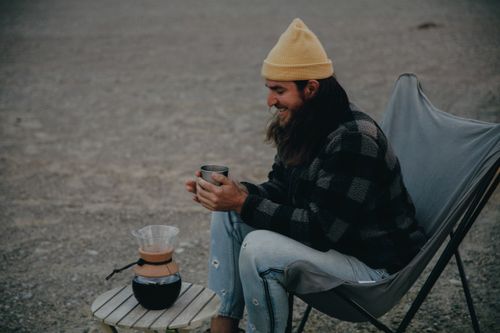 Free A relaxed man in a checkered coat with a yellow beanie enjoys coffee outdoors, embodying a rustic lifestyle. Stock Photo