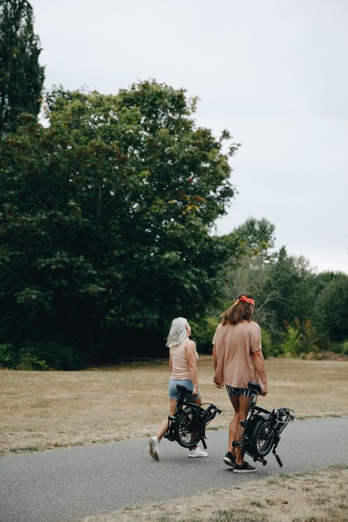 Free A couple walking with folding bicycles on a park path surrounded by trees. Stock Photo