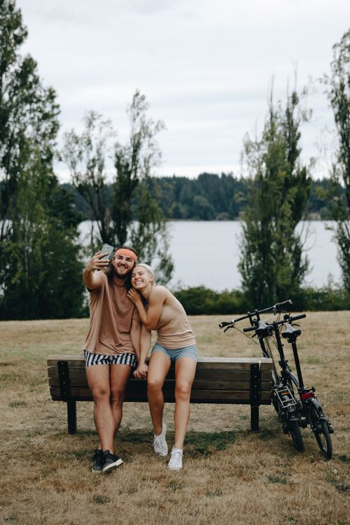 Free Happy couple sitting on a bench taking a selfie by the lake with an electric bike nearby. Outdoors, summer fun. Stock Photo