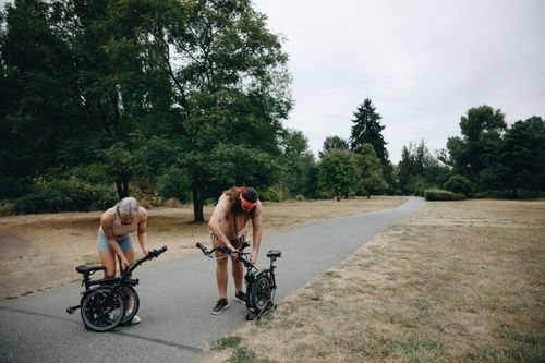 Free A couple adjusting their bicycles on a park pathway during a cloudy day, enjoying an outdoor activity. Stock Photo